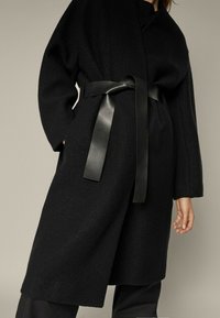 Sort oversized frakke lavet af tekstureret stof, med en omslaget design og et bredt sort læderbælte i taljen. Lange ærmer strækker sig til midt på underarmen.