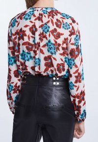 Blouse fleurie avec des fleurs rouges et turquoise sur un fond blanc, manches longues, encolure froncée ; associée à un pantalon en cuir noir taille haute avec des détails à boutons-pression.