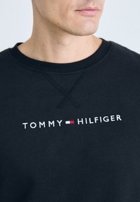 Sudadera de algodón negra con cuello redondo, que presenta el logo bordado "TOMMY HILFIGER" y un pequeño emblema en rojo, blanco y azul.