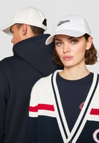 Hvit baseballcap med svart broderi, marineblå hettegenser og stripete cardigan i marineblått, hvitt og rødt. Strukturerte stoffer synlige.