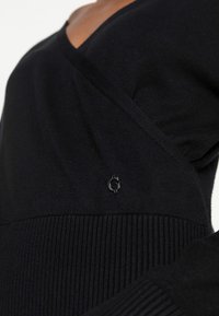 Top noir en maille avec un décolleté en V profond et des accents côtelés. Présente un petit logo en relief sur le côté, offrant une texture lisse.