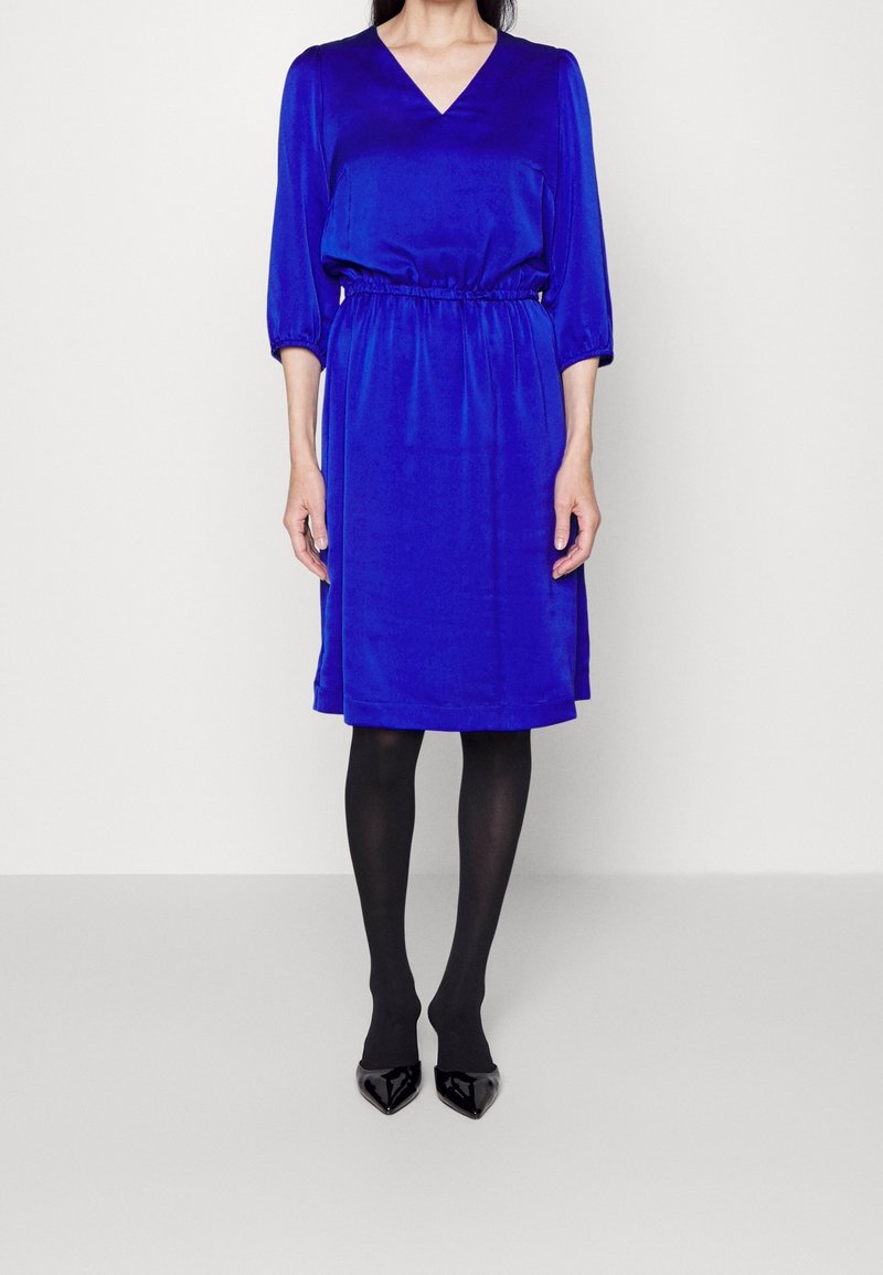 Vestido de satén azul con escote en V, mangas 3/4, cintura elástica, largo hasta la rodilla, combinado con medias negras y tacones de charol puntiagudos.