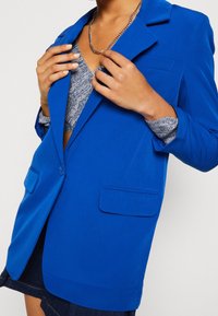 Blå blazer i slät tyg med slagkläppar, två framfickor och en enkel knäppning. Lager över en mönstrad topp.