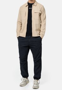 Homme portant une veste beige zippée sur une chemise noire, pantalon bleu marine foncé et baskets claires, debout les mains dans les poches.