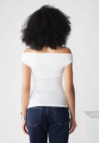Witte off-shoulder top met een aansluitende pasvorm, gladde textuur en een geplooid effect, gecombineerd met donkerblauwe denim jeans met achterzakken.