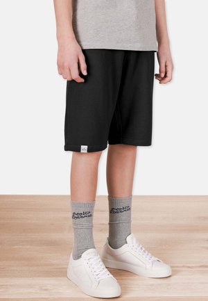 Sorte bomuldsshorts med elastisk talje og en lille logo-patch. Kombineret med grå ankelstrømper og hvide sneakers. Glat tekstur.