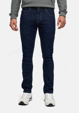 Mężczyzna ubrany w ciemnoniebieskie dopasowane dżinsy, szarą bluzę w paski z długim rękawem oraz białe trampki, stojący na białym tle.