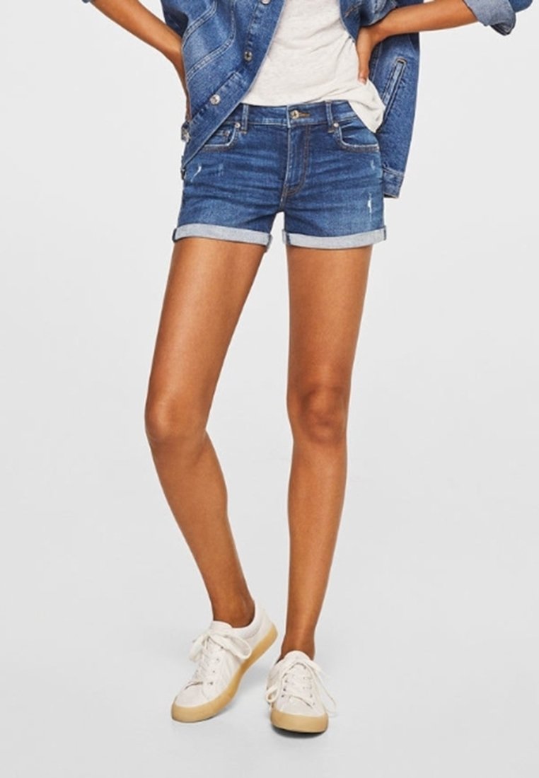 Jeanshort in een donkerblauwe wassing met omgeslagen zomen. Gekenmerkt door een grijs shirt en witte sneakers, wat een casual zomerse stijl laat zien.
