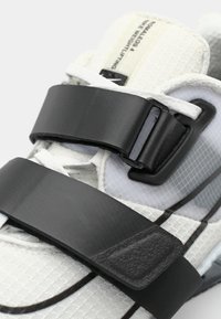 Chaussures de musculation blanches avec une tige texturée, des bandes velcro noires, des accents argentés et une semelle renforcée pour plus de stabilité.