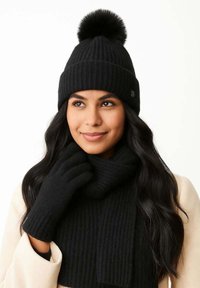 Sieviete ar melnu adītu cepuri ar pom-pom, atbilstošu šalli un cimdiem, ar gariem tumšiem matiem un gaišas krāsas mēteli.