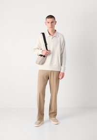 Beiger Pullover mit Kragen, hellbraune Hose und beiger Sneakers; trägt eine kleine graue Umhängetasche. Glatter Stoff, lockere Passform.