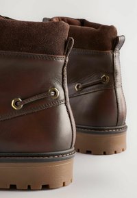 Botas cortas de cuero marrón con un puño de ante, ojales en tono dorado y suelas de goma duraderas que presentan un patrón de agarre para tracción.