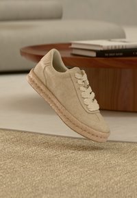 Μπεζ υφασμάτινο αθλητικό παπούτσι με χοντρή πλεκτή σόλα και κορδόνια στο ίδιο χρώμα, που αιωρείται πάνω από ένα υφασμάτινο μπεζ χαλί σε ένα μοντέρνο σαλόνι.