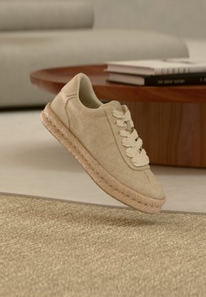 Baskets en tissu beige avec une semelle épaisse tressée et des lacets assortis, flottant au-dessus d'un tapis beige texturé dans un salon moderne.