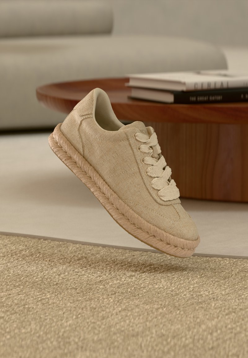 Μπεζ υφασμάτινο αθλητικό παπούτσι με χοντρή πλεκτή σόλα και κορδόνια στο ίδιο χρώμα, που αιωρείται πάνω από ένα υφασμάτινο μπεζ χαλί σε ένα μοντέρνο σαλόνι.