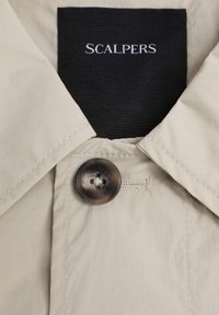 Jachetă bej cu guler ascuțit, un nasture maro și un etichetă cu "SCALPERS" în text alb pe un fundal negru. Textură fină.