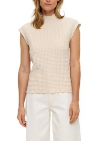 Beige textuur top met een hoge hals en korte mouwen, met een gegolfde zoom. Draag met witte broeken, die het patroon van de stof accentueren.