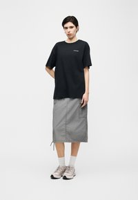 Νέος άνθρωπος στέκεται μπροστά σε λευκό φόντο φορώντας μαύρο t-shirt Columbia, γκρι midi cargo φούστα, λευκές κάλτσες και ροζ αθλητικά παπούτσια.