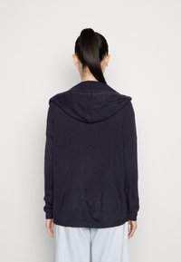 Tmavě námořnická pletená mikina s kapucí s žebrovaným límcem a manžetami, se středovým švem na zádech a volnějším střihem. Měkká textura.