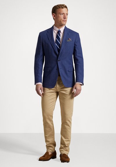 Blazer de lino azul, camisa rosa claro con rayas azules, pantalones beige y mocasines marrones. El pañuelo de bolsillo añade un detalle de color.