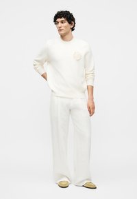 Jeune homme aux cheveux bouclés portant un pull écru avec un détail floral, un pantalon blanc à jambes larges et des chaussures beige, debout devant un fond uni.