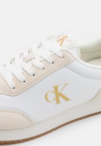 Bijela tenisica s naglascima od svijetlosmeđeg sueda, koja ima teksturiranu površinu, bijele vezice i zlatni "ck" logo sa strane.