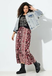 Mujer con cabello rizado que lleva una chaqueta de mezclilla clara, una camiseta gráfica negra, una falda midi de estampado rojo y botas de combate negras, posando contra una pared blanca.
