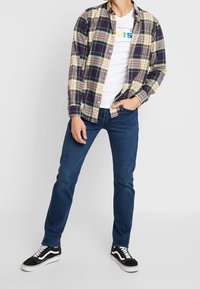 Rutigt flanellskjorta i mörk lila, gröna och gula toner, bärs över en vit t-shirt. Blå jeans och svarta sneakers fullbordar looken.