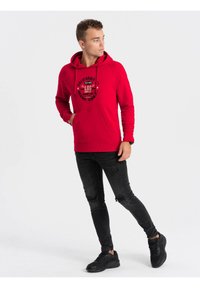 Rode hoodie met een voorkruiszak, voorzien van een zwart grafisch ontwerp. Gecombineerd met zwarte versleten jeans en zwarte sneakers.