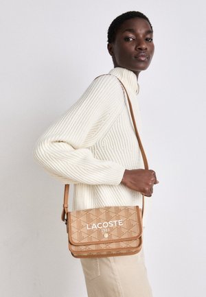 Kobieta w białym, prążkowanym swetrze, nosząca na ramieniu beżową, wzorzystą torebkę na ramię Lacoste z zapięciem na guzik.