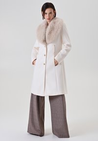 Cappotto in lana crema con un ampio colletto in pelliccia sintetica, design doppiopetto, bottoni dorati e tasche laterali, abbinato a pantaloni a gamba larga a quadri.