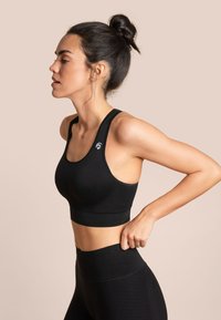 Schwarzer Sport-BH mit enganliegendem Design, breiten Trägern und glatter Textur. Mit markiertem Logo auf der linken Schulter. Kombiniert mit hochgeschnittenen, schwarzen Leggings.