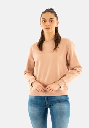 Sudadera color rosa claro con cuello redondo, puños acanalados y cintura elástica. Presenta un logotipo de la marca en relieve en el pecho. Combinada con jeans azules.