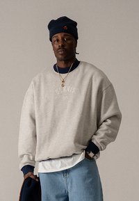 Szürke, kerek nyakkivágású pulóver hímzett "BLK VIS NY" logóval, tengerészkék részletekkel a mandzsettákon, fehér ing fölött viselve, világoskék farmerral párosítva.