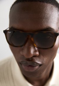 Ruskeastiilon aurinkolasit tummilla linsseillä. Kehyksissä Ray-Ban -logo. Lähikuva korostaa muotoa ja rakennetta.