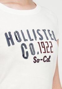 Hvit bomull T-skjorte med brodert marineblå og burgunder tekst: "HOLLISTER CO. 1922 So-Cal." Klassisk rund hals og figursydd ermedesign.