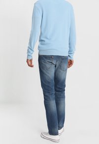 Man som bär en ljusblå tröja, urblekta blå jeans och vita sneakers, står med ryggen mot kameran framför en enkel bakgrund.