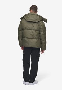 Olive green puffer jacket with a hood, boxy design, quilted sections, and back straps. Worn with black cargo pants and white sneakers.