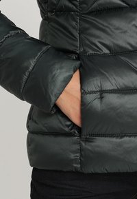 Main dans la poche d'une doudoune noire matelassée avec des coutures visibles et un tissu légèrement brillant.