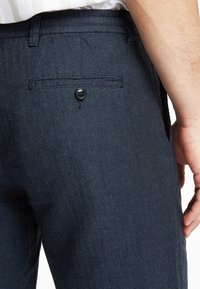 Kalhoty z námořnického modrého herringbone vzoru s jednou zadní kapsou uzavíratelnou černým knoflíkem. Hladká textura a přiléhavý střih.