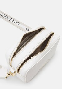 Bolsa transversal sintética branca com fechos duplos, hardware dourado e acabamento texturizado. Inclui uma alça de tecido com o logótipo "Valentino".