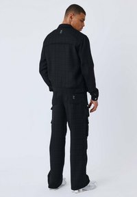 Giacca nera texturizzata e pantaloni cargo con tasche, caratterizzati da un motivo sottile. Accenti in metallo argentato e un dettaglio del logo sulla giacca.