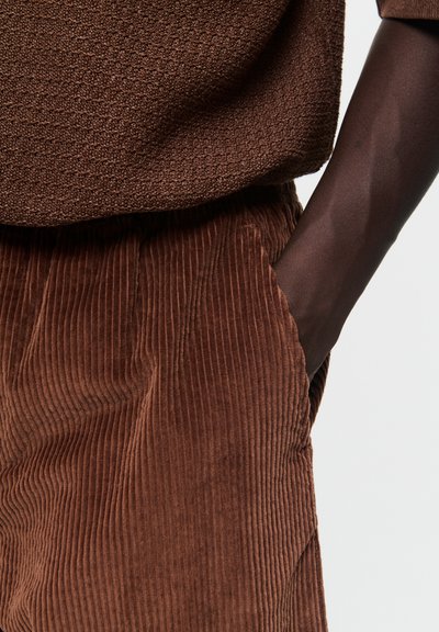 Un pull en laine marron texturé assorti à un pantalon en velours côtelé. Le pull présente un motif tissé ; le pantalon, quant à lui, est doté de rainures verticales, créant une riche apparence tactile.