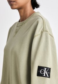 Felpa verde oliva con scollatura rotonda e bordi a costine. Presenta un'etichetta in tessuto nera con "Calvin Klein Jeans" sulla manica sinistra.
