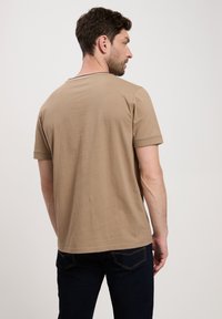 Koszulka z krótkim rękawem w kolorze beżowym, wykonana z bawełny, z okrągłym dekoltem i prostym wzorem, noszona z ciemnoniebieskimi dżinsami.