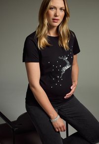 Svart t-shirt med en glittrande silverren design och stjärnor. Modellen håller sin gravida mage, iförd mörka jeans och ett armband.