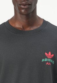 Maglietta nera in cotone con collo rotondo, caratterizzata da un logo grafico Adidas rosso e verde sul lato sinistro. Texture liscia.