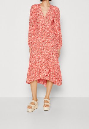 Femme portant une robe midi rouge à fleurs, manches longues, col en V, avec un ourlet à volant et des sandales compensées beiges, debout devant un fond uni.