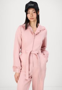 Macacão rosa com colarinho e punhos franzidos, fecho na frente, cintura com cinto e bolsos laterais. Acompanhado de um boné preto com o texto "Juicy".