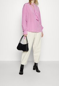 Blusa rosa a maniche lunghe con pannello arricciato, abbinata a pantaloni crema. Tiene una borsa nera e indossa stivaletti neri lucidi.